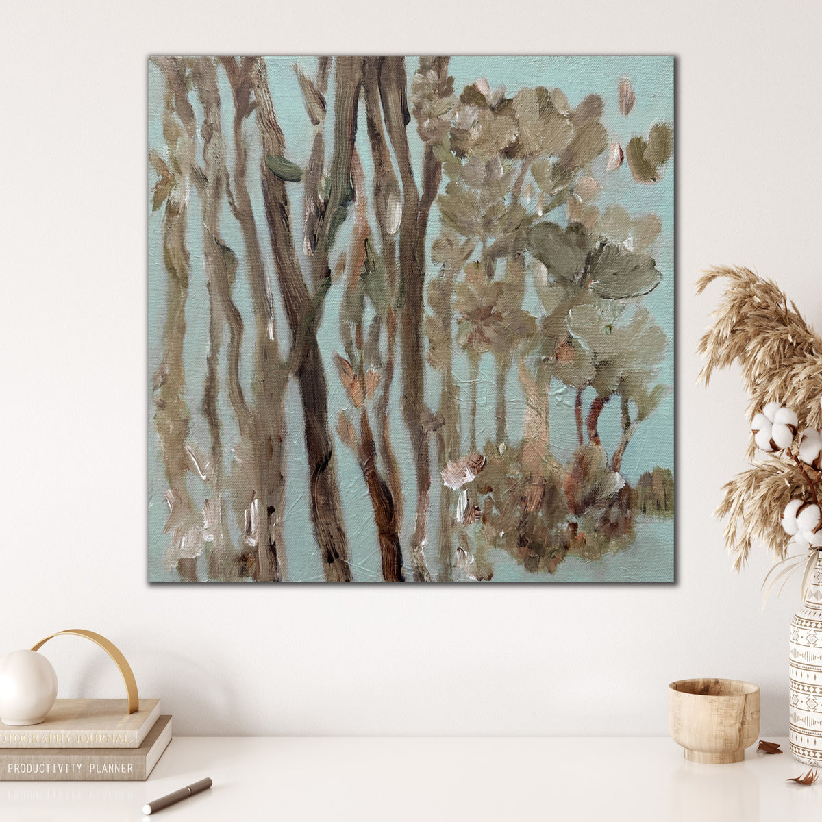 Square painting of nature tree scape with soft blue background and tree trunks and foliage in natural colours of greens and browns hanging on white wall in modern interior.