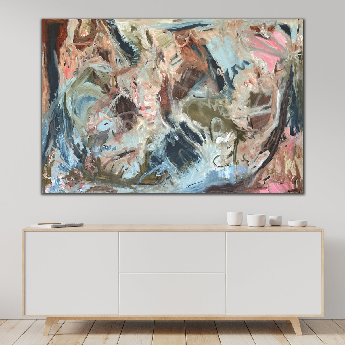 A large, contemporary abstract painting in shades of blue, brown, pink and green, hanging on wall above sideboard in modern home. 